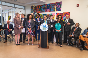 Eine Gruppe von Menschen steht vor einem Podium mit einem Mikrofon und einer Tafel mit der Aufschrift "Sancho-Stadtrat tritt dem Stadtrat bei", während andere im Hintergrund sitzen und stehen, Flaggen, eine Tafel, ein Feuerlöscher und Deckenleuchten an der Rückwand.
