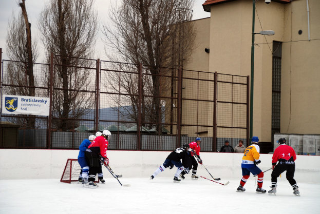 Menschen beim Eisschnelllauf auf einer Eisbahn mit Gebäuden, Bäumen, einer Straßenlaterne, einem Namensschild und Zäunen im Hintergrund unter dem Himmel.