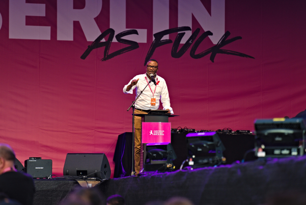 Ein Mann in weißem Hemd und braunen Hosen steht auf einer Bühne und hält ein Mikrofon in der Hand, mit einem pinken Banner mit der Aufschrift 'Berlin as Fuck' im Hintergrund und einer Menge davor.
