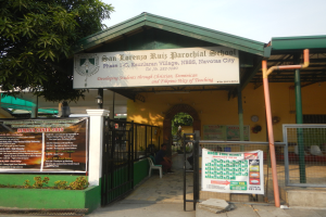 Der Eingang der San Lorenzo Ruiz Pfarrschule hat eine Tafel mit Text, einen Metallzaun, Menschen auf Stühlen unter einem Dach, Pfosten, ein Schild, Pflanzen, einen Baum und einen bewölkten Himmel.