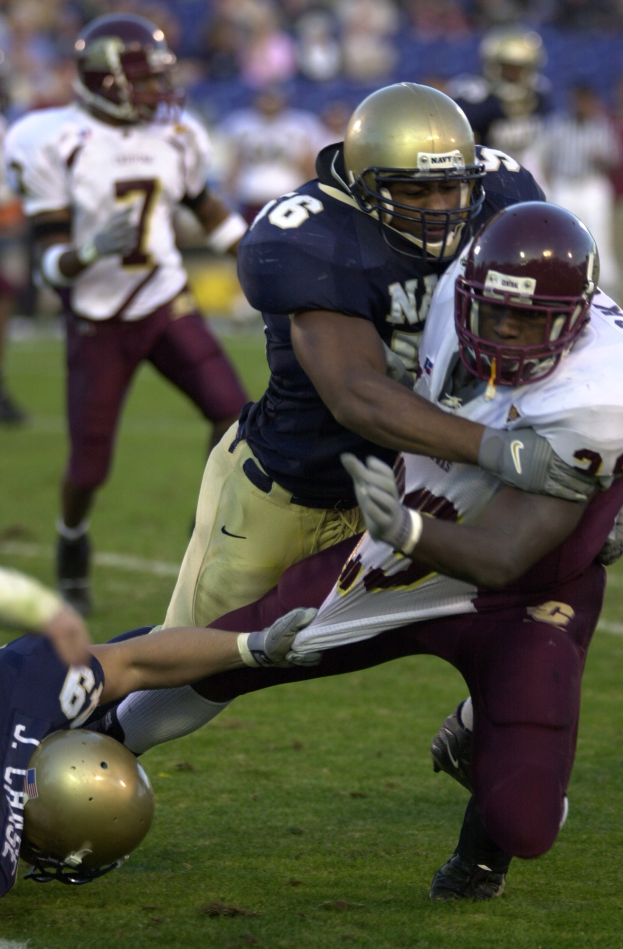 Zwei Fußballspieler in Helmen und Uniformen kämpfen auf dem Feld gegeneinander, während andere im Hintergrund zusehen.
