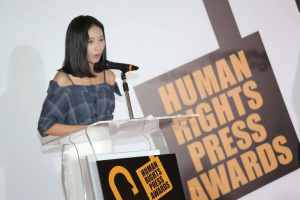 Eine Frau in einem schwarzen Kleid steht an einem Podium mit einem Mikrofon und hält eine Rede oder Präsentation vor einem Banner, auf dem "Human Rights Press Awards" steht. Ihr Haar ist zurückgebunden und ihr Gesichtsausdruck ist ernst und konzentriert.
