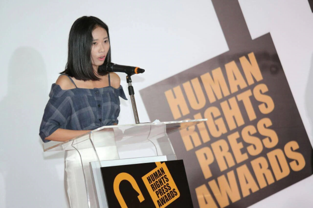 Eine Frau in einem schwarzen Kleid steht an einem Podium mit einem Mikrofon und hält eine Rede oder Präsentation vor einem Banner, auf dem "Human Rights Press Awards" steht. Ihr Haar ist zurückgebunden und ihr Gesichtsausdruck ist ernst und konzentriert.