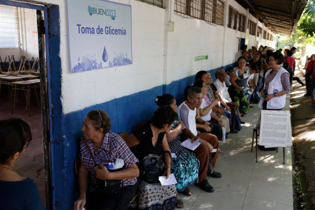 Eine Gruppe von Menschen sitzt auf Bänken vor einem Gebäude mit einer Texttafel an der Wand, einer Tür links und Bäumen im Hintergrund, die auf die Stimmabgabe an einer Wahllokal in Guadalajara, Mexiko, warten.