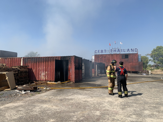 Zwei Feuerwehrleute in Helmen vor einem rauchenden Gebäude mit einer Rohrleitung, umgeben von Schutt, Containern und einem Namensschild, vor einem Hintergrund aus Bäumen und einem klaren Himmel.