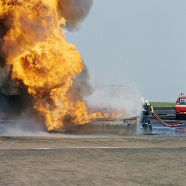 Feuerwehrlöschfahrzeug in Flammen an der Stra√enseite mit zwei Helmträgern, die Schläuche halten, einem Fahrzeug im Hintergrund und dem Himmel.