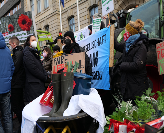 Eine Gruppe maskierter Menschen mit Protestschildern vor einem Lastwagen, mit einem Tisch voller Gegenstände, Topfpflanzen und einem Gebäude mit Fahne im Hintergrund.