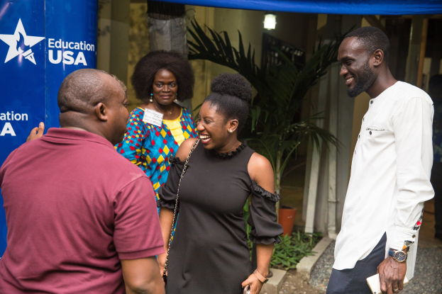 Gruppe von Menschen, die vor einem blauen Zelt mit einem Banner, Topfpflanzen, einer Säule, einer Wand und Deckenleuchten sprechen.