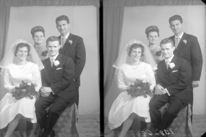 Ein Schwarz-Weiß-Hochzeitsfoto einer Braut in einem weißen Kleid und Schleier, die einen Strauß hält, mit einem Bräutigam dahinter, der strahlt, und zwei anderen Menschen dahinter, die ebenfalls strahlen, mit einem Vorhang und einer Wand im Hintergrund.