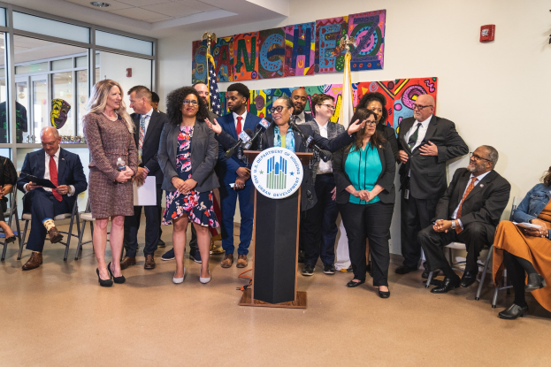 Gruppe von Menschen vor einem Rednerpult mit einem Mikrofon und einer Tafel mit dem Text "Sancho City Councilman Joins City Council", mit sitzenden und stehenden Teilnehmern, Flaggen, einem Feuerlöscher und Deckenleuchten im Hintergrund.