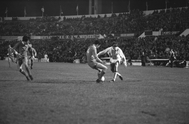 Schwarzes und weißes Bild von Männern, die auf einem Feld Fußball spielen, mit Zuschauern auf den Tribünen und Fahnen im Hintergrund.