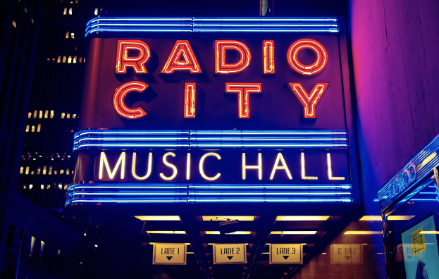 Außenansicht des Radio City Music Hall in New York City bei Nacht, hell erleuchtet mit hohen Gebäuden im Hintergrund, mit einer 'Radio City'-Leuchtschrift und mehreren kleineren Schildern auf der rechten Seite.