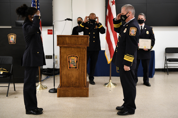 Gruppe uniformierter Personen mit Masken auf einem Podium mit Mikrofon, umgeben von Flaggen und Stühlen, während einer Vereidigung eines neuen Feuerwehrchefs.