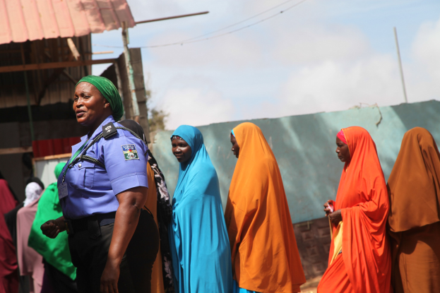 Eine Gruppe von Frauen, die vor einem Gebäude mit einem Polizisten im Vordergrund und einer Kulisse aus Baracken, Strommasten, Bäumen und einem klaren blauen Himmel steht.