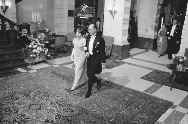 Schwarz-weißes Foto einer Braut und eines Bräutigams, die durch die Lobby eines Hotels gehen, umgeben von Gästen, einem Tisch, Stühlen, Blumenarrangements, Lampen, einer Treppe und einem Geländer.