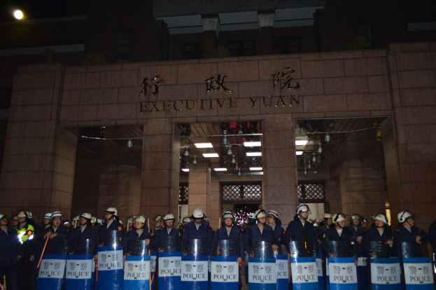 Gruppe von Polizisten in Helmen und Schilden vor einem Gebäude mit beleuchteter Wand und Decke.