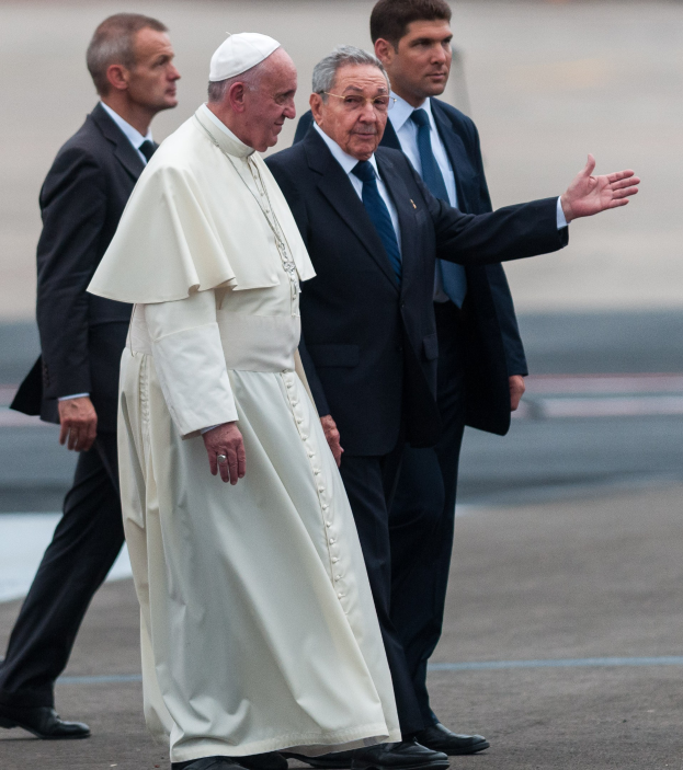 Papst Franziskus in einem weißen Gewand und einer Mütze, begleitet von vier Männern in schwarzen Blazern und Krawatten, beim Ankunft an einem Flughafen im Petersplatz im Vatikan.