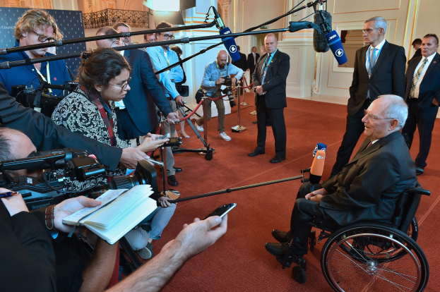 Ein Mann im Rollstuhl in Anzug und Krawatte von Reportern mit Kameras und Mikrofonen umgeben.