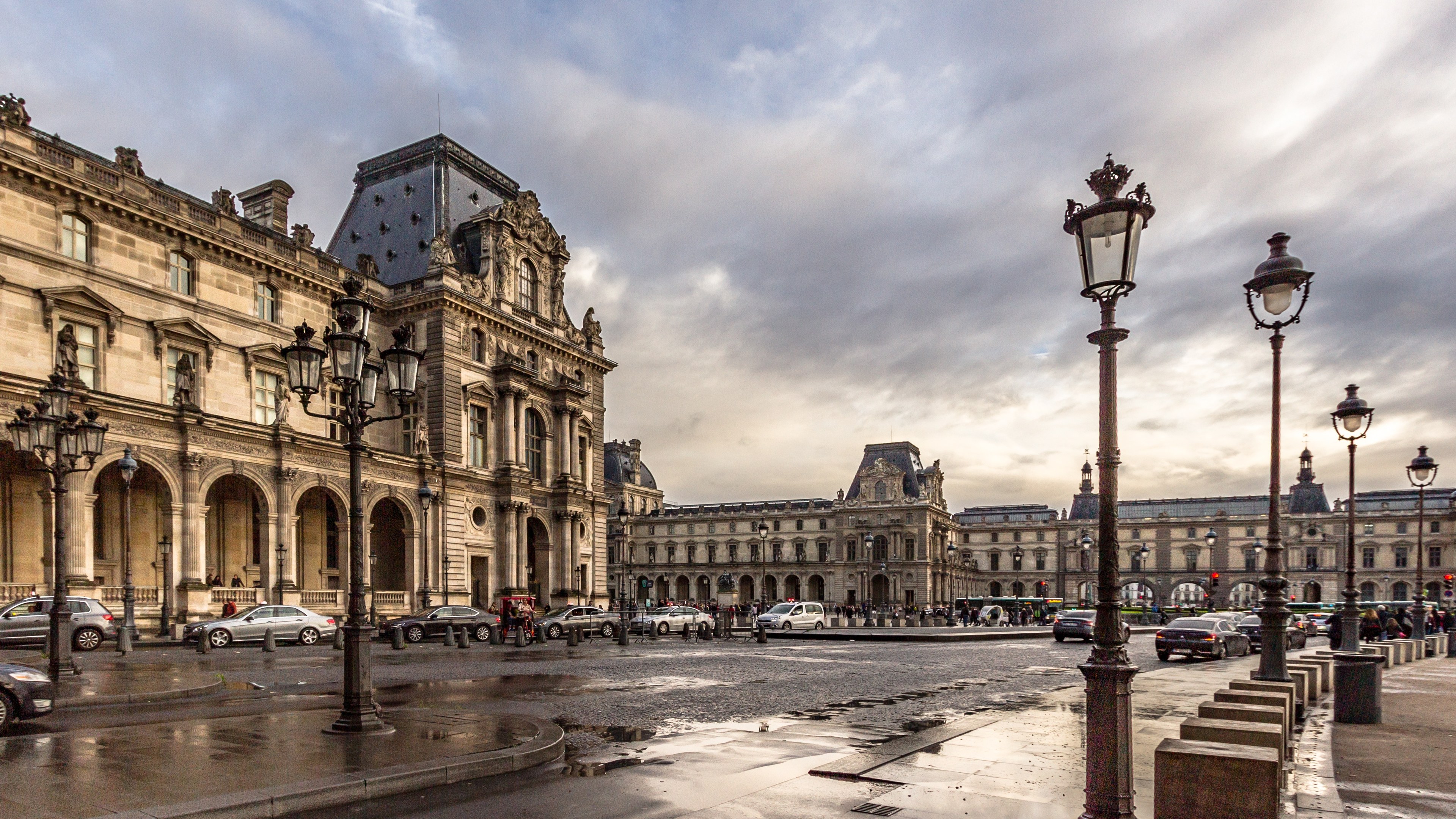 Außenansicht des Louvre-Museums in Paris mit seiner ikonischen Architektur, Straßenlaternen, Lichtern, Fahrzeugen, Passanten und einem bewölkten Himmel.