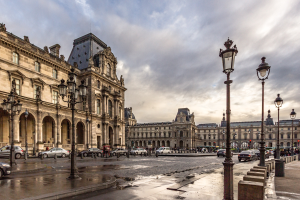 Außenansicht des Louvre-Museums in Paris mit seiner ikonischen Architektur, Straßenlaternen, Lichtern, Fahrzeugen, Passanten und einem bewölkten Himmel.