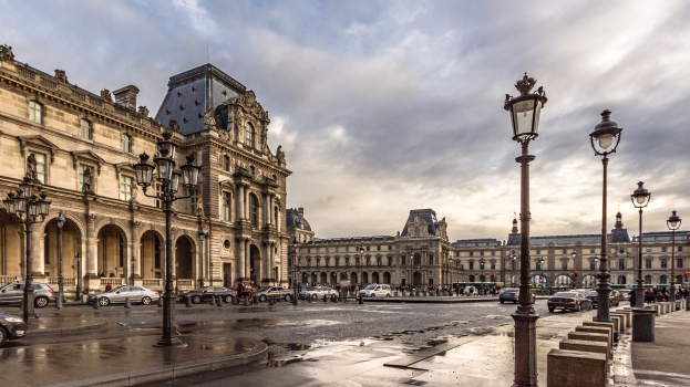 Außenansicht des Louvre-Museums in Paris mit seiner ikonischen Architektur, Straßenlaternen, Lichtern, Fahrzeugen, Passanten und einem bewölkten Himmel.