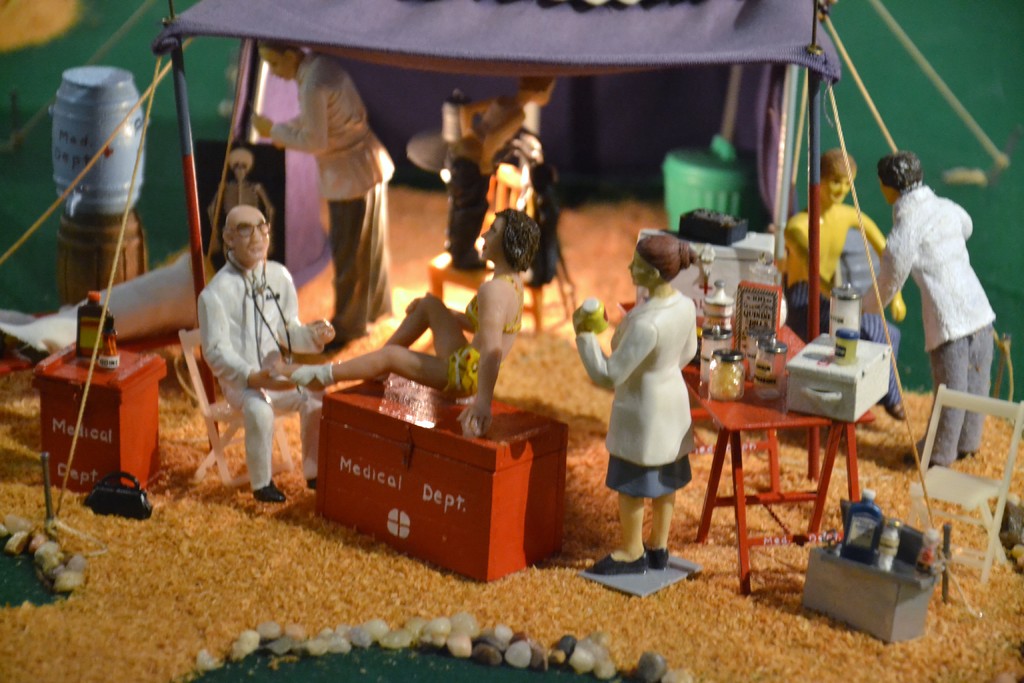 Eine Puppe steht auf einer Box mit der Aufschrift 'medizinisches Dept' mit einem Tisch voller Gegenstände daneben, alles unter einem violetten Zelt, mit zusätzlichen Gegenständen im Hintergrund.