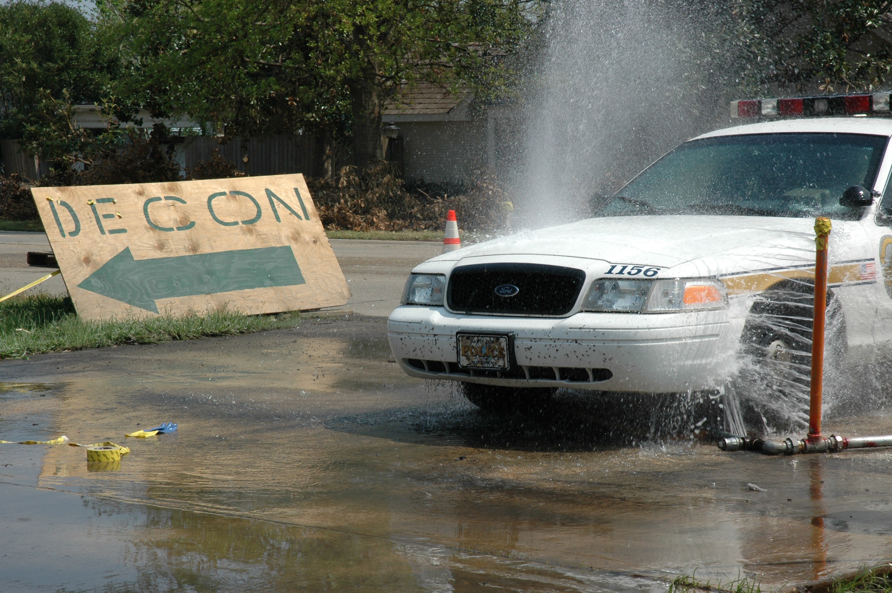 Ein Polizeiwagen fährt durch eine Pfütze, mit einem Schild, einer Verkehrskegel und anderen Gegenständen auf dem Boden, vor einem Hintergrund aus Bäumen, Gebäuden und einem klaren Himmel.