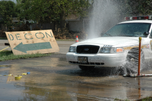 Ein Polizeiwagen fährt durch eine Pfütze, mit einem Schild, einer Verkehrskegel und anderen Gegenständen auf dem Boden, vor einem Hintergrund aus Bäumen, Gebäuden und einem klaren Himmel.