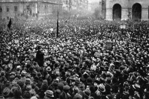 Ein Schwarz-Weiß-Bild einer großen Menschenmenge vor einem Gebäude, mit einer Person in der Mitte, die eine Fahne und ein Schild hält, wahrscheinlich bei einer Suffragetten-Demonstration.