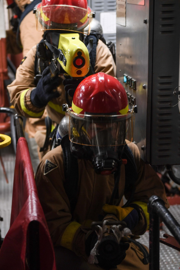 Feuerwehrleute in Schutzausrüstung, einer hält einen Feuerlöscher, mit einem metallischen Objekt und einem roten Tuch im Hintergrund.