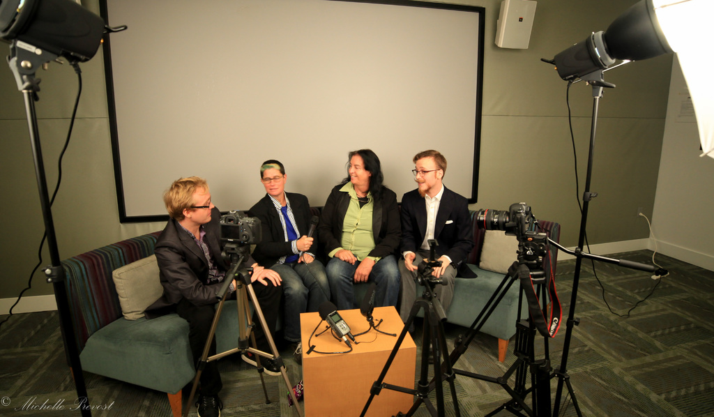 Eine Gruppe von drei Personen sitzt auf einem Sofa und gibt ein Interview, wobei eine Person in einem schwarzen Mantel und Hose links, eine Person in einem Mantel und einer blauen Krawatte in der Mitte mit einem Mikrofon und eine Person rechts mit einem Lächeln zu sehen ist. Ein Mikrofonrecorder befindet sich auf dem mittigen Tisch, Kameras sind auf der rechten und linken Seite und große Lichtstative sind vorhanden, mit einem Projektionsschirm im Hintergrund.