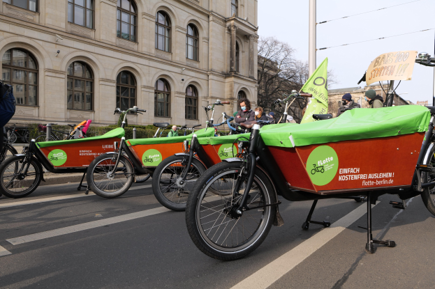 Eine Gruppe von Fahrrädern, die entlang einer Straße geparkt sind, mit einer Person in der Nähe, vor Gebäuden und Bäumen unter einem klaren Himmel, Teil einer Fahrrad-Sharing-Kampagne.
