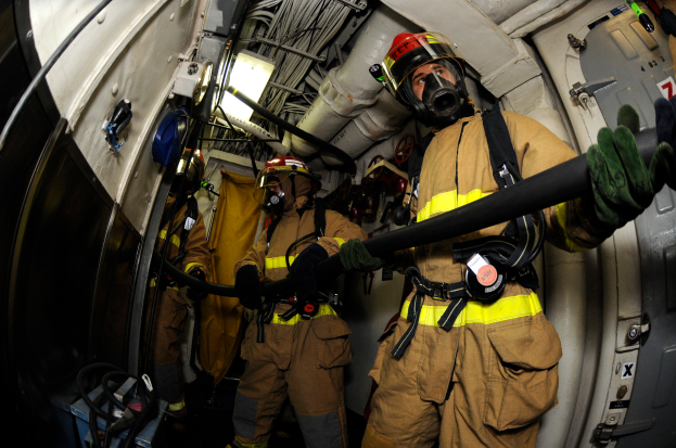 Eine Gruppe von Feuerwehrleuten in Helmen und Handschuhen hält einen Schlauch an der Rückseite eines Flugzeugs, mit Kabeln, Lampen und anderen Gegenständen im Hintergrund und Text am unteren Bildrand.
