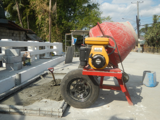 Betonmischer auf einem Zementlaster auf der Straße, mit Betonblöcken, einem Eimer, einer Karre, einem Zaun, einem Haus, Bäumen, Strommasten mit Drähten und einem bewölkten Himmel.