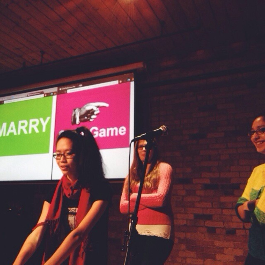 Drei Frauen, die nach links schauen, ein Mikrofonständer in der Mitte, eine Wand im Hintergrund und ein Bildschirm mit Text auf der linken Seite.