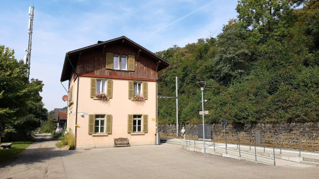 Ferienhaus zum Verkauf in Baden-Württemberg, Deutschland, mit einem Gebäude mit vielen Fenstern, einem Vorgartenzaun, Straßenmöbeln, Grünfläche und einem bewölkten Himmel.