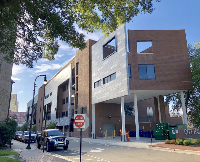 Großes mehrstöckiges Stadtzentrumsgebäude mit zahlreichen Fenstern, Straßeninfrastruktur einschließlich Pfosten, Lampen und Verkehrszeichen, umgeben von Fahrzeugen, Grünflächen und einem bewölkten Himmel.
