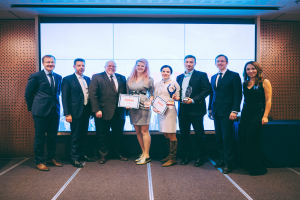 Eine Gruppe strahlender Menschen hält Zertifikate und steht vor einem Bildschirm, mit einem rechts daneben stehenden Tisch mit schwarzem Tuch und einem Gegenstand an der Wand im Hintergrund, feiert ihren Sieg bei den International Business Awards 2018.