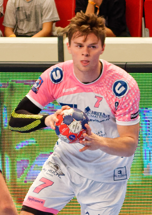 Ein Mann in einem pinken Hemd hält einen Fußball in der Hand, während im Hintergrund Menschen auf Stühlen sitzen.