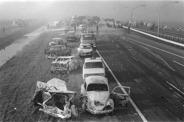 Ein Schwarz-Weiß-Foto von mehreren verunglückten Autos am Straßenrand bei Nacht, mit Laternen, nahestehenden Menschen, Bäumen und einem Gewässer im Hintergrund.