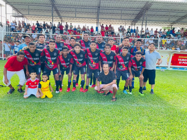 Gruppe von Männern, die auf einem Fußballfeld mit einem "Fotos Ugarte U-17 vs U-18"-Banner feiern, umgeben von Metallzaun, Pfählen, einem Dach und einem bewölkten Himmel.