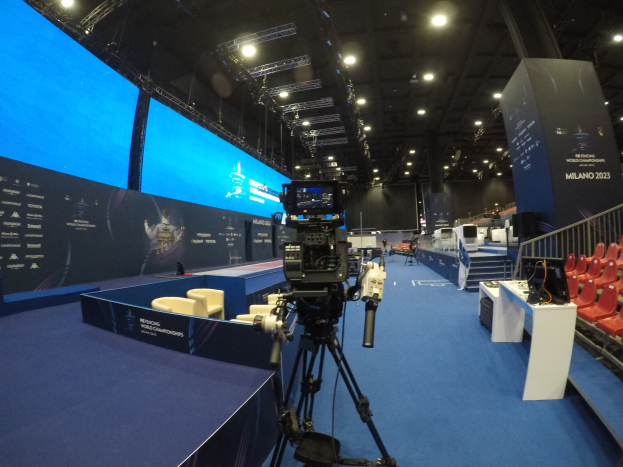 Eine Videokamera auf einem Stativ vor einem großen Bildschirm auf der Interchange World Championships-Veranstaltung positioniert, mit Sitzplätzen, Bannern und Beleuchtung.