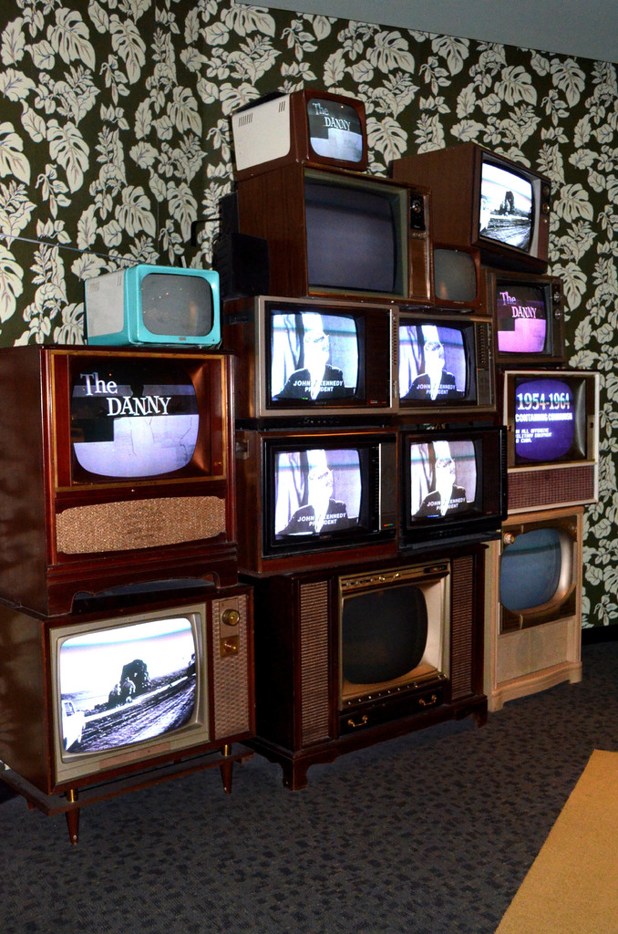 Zwei Fernseher stehen nebeneinander auf einer Oberfläche, mit einem Blumenmuster-Vorhang im Hintergrund und einem Boden, der unten im Bild sichtbar ist.