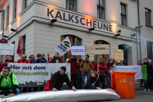 Eine Gruppe von Menschen steht vor einem Gebäude und hält Schilder und Plakate hoch, während zwei Personen im Vordergrund sitzen und ein Müllcontainer rechts zu sehen ist, bei einer Demonstration in Deutschland, mit Gebäuden, Fenstern, Lichtern und Schildern im Hintergrund.