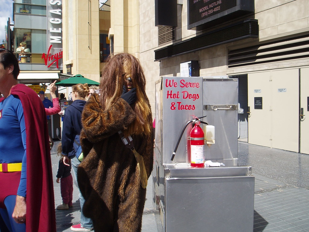 Eine Menschenmenge, darunter einige in Kostümen, versammelte sich vor Gebäuden mit Schildern, einer Box mit Aufschrift und einem Feuerlöscher.