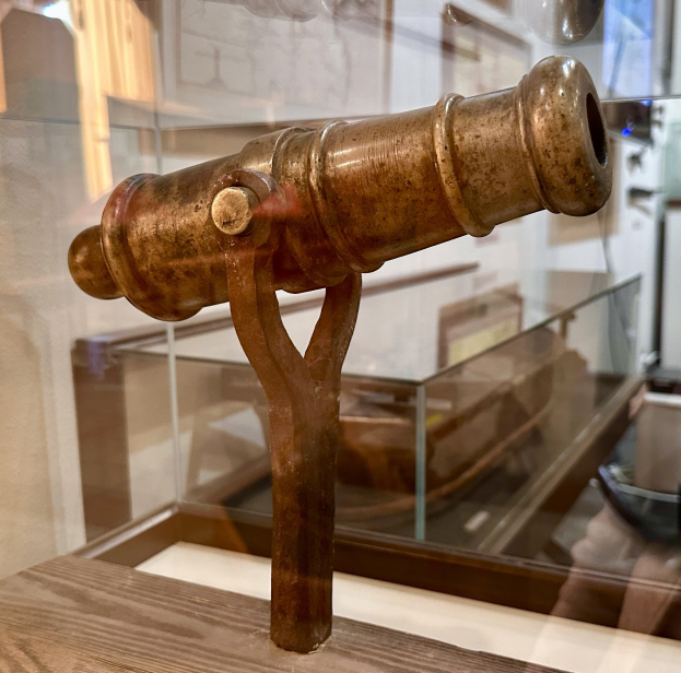 Alte Messingkanone in einer Glasvitrine mit einem hölzernen Unterbau, vor einem Hintergrund mit gerahmten Wandausstellungen in einem Museums Setting.