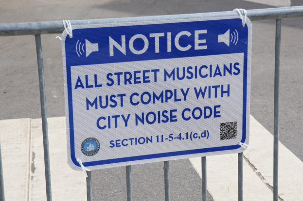 Anschlagtafel an einem Zaun mit einem Schild, auf dem "Hinweis: Alle Straßenmusiker müssen die städtische Lärmvorschrift einhalten" steht und eine Straße im Hintergrund.