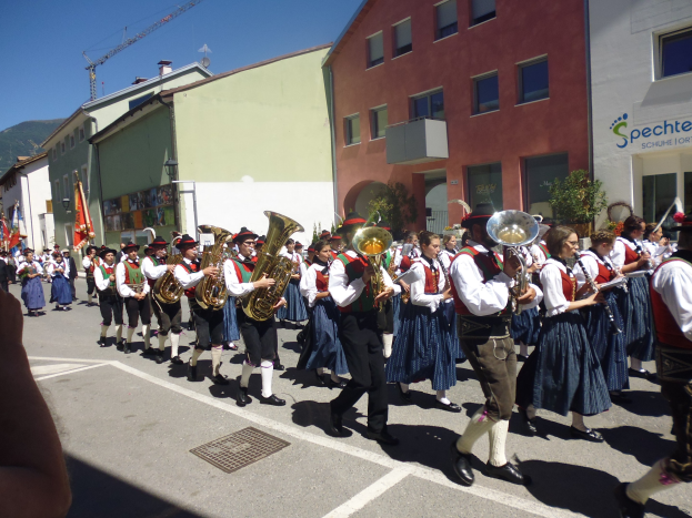 Eine Gruppe von Menschen in traditioneller bayrischer Tracht, die Musikinstrumente spielen, während sie eine von Gebäuden gesäumte Straße entlanggehen, einige halten Fahnen, mit einem Hügel und einem klaren blauen Himmel im Hintergrund.