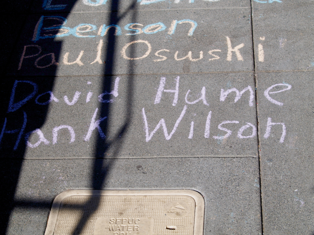 Ein Gehweg mit Kreide-Schrift, die wie ein Denkmal aussieht, ein Gullideckel am Boden und ein Schatten eines Baums auf der linken Seite.