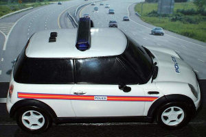 Ein weißes Polizeiauto ist im Vordergrund geparkt, mit vielen Autos, die sich auf der Straße dahinter bewegen, umgeben von Gras und Bäumen auf beiden Seiten, in einer ländlichen Umgebung.